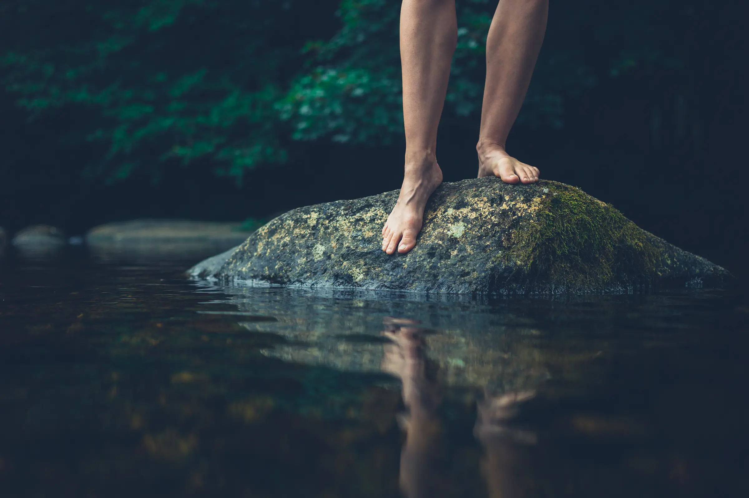 female legs and feet standing on a boulder just above the waterline in a creek